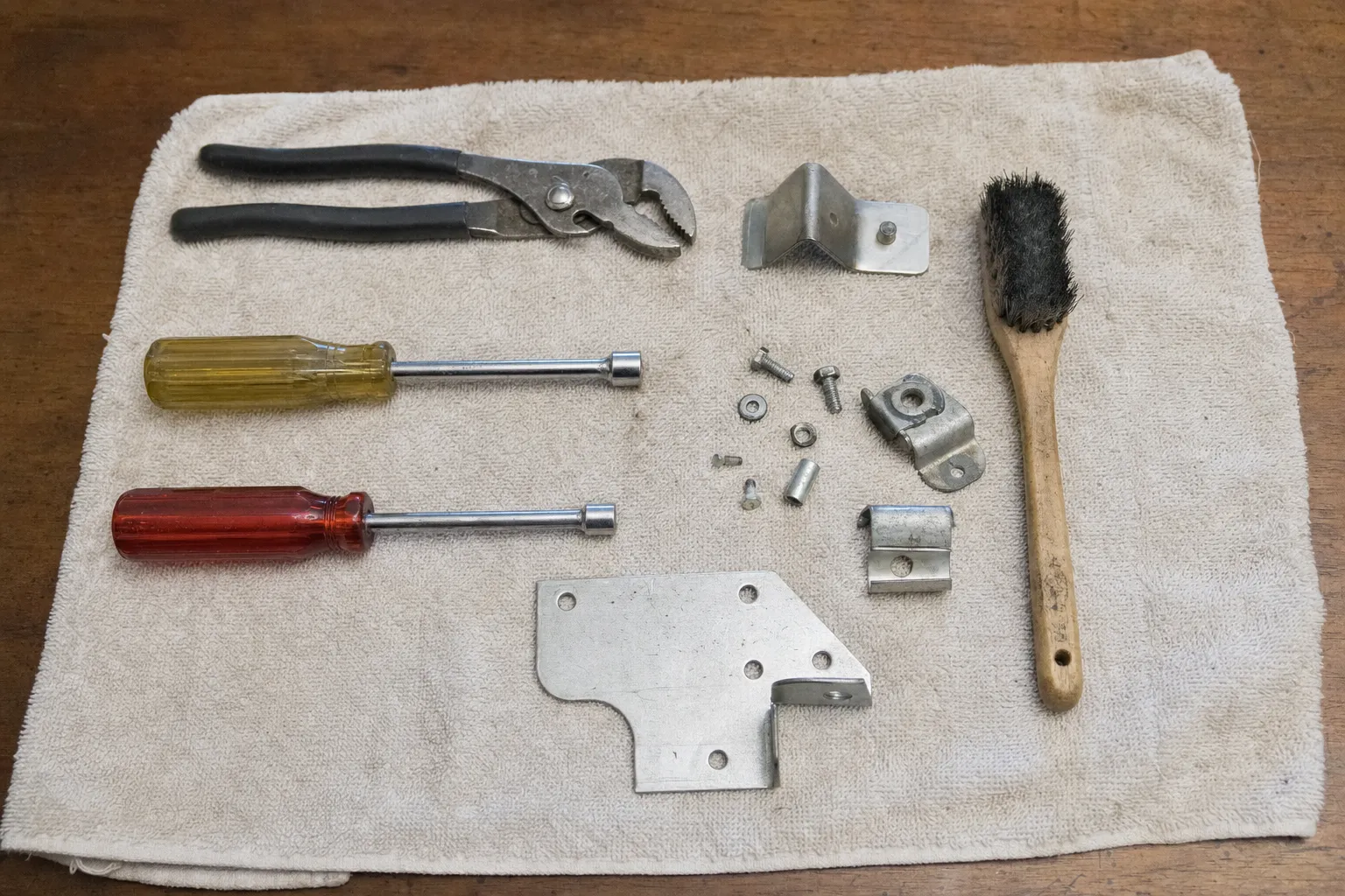 Slip-joint pliers, two nut drivers and a wire brush on a shop towel beside brackets and fasteners